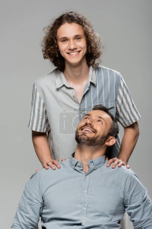 positive father smiling and looking at happy teenage son isolated on grey 