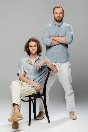 full length of good looking man standing with folded arms near teenage son on grey 