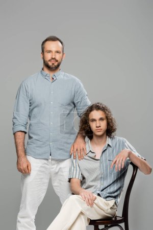 good looking dad standing next to teenage son with curly hair isolated on grey 