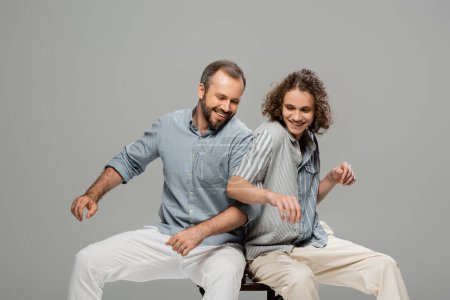 happy father and son having fun and pushing each other while sitting on one chair isolated on grey 