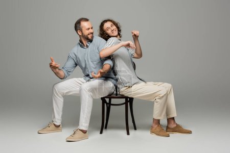 Smiling father and teenage son having fun and pushing each other while sitting on same chair on grey