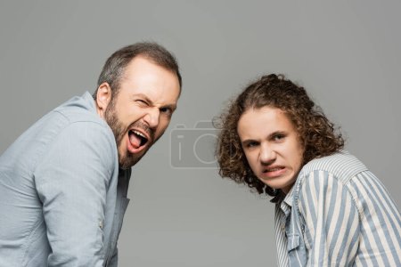 father with opened mouth and disgusted teenage son grimacing and looking at camera isolated on grey 
