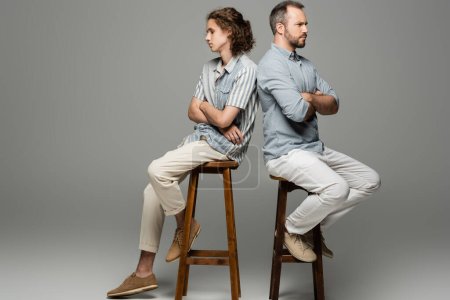 beleidigter Vater und Teenager-Sohn sitzen mit verschränkten Armen Rücken an Rücken auf grau 