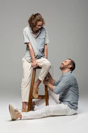 curly teenage guy smiling while sitting on high chair next to happy dad on grey 