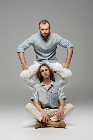 powerful and bearded dad sitting on chair next to curly teenager boy on grey 