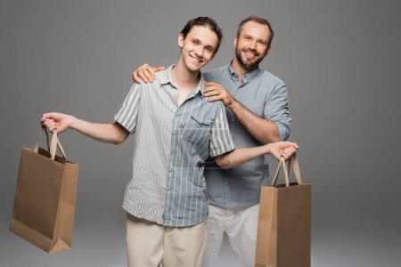 glücklicher Teenager mit Einkaufstüten neben fröhlichem Papa auf grau 