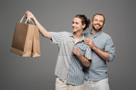 happy teenage boy holding shopping bags near positive father isolated on grey 