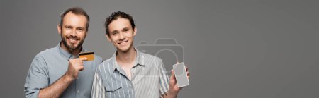 happy father holding credit card next to teenage son with smartphone in hand isolated on grey, banner 