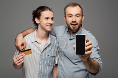 amazed man holding smartphone with blank screen next to teenage son with credit card in hand isolated on grey 