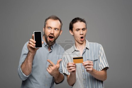 Foto de Asombrado padre sosteniendo teléfono inteligente con pantalla en blanco junto a hijo adolescente conmocionado con tarjeta de crédito en la mano aislado en gris - Imagen libre de derechos