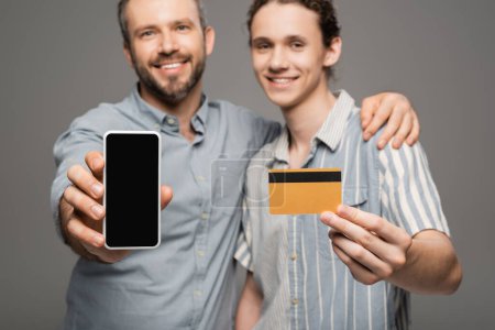 overjoyed father holding smartphone with blank screen next to happy teenage son with credit card in hand isolated on grey 