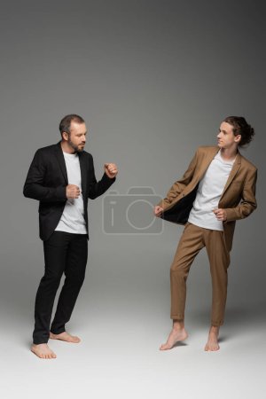full length of well dressed father dancing with teenage and barefoot son in suit while having fun on grey 