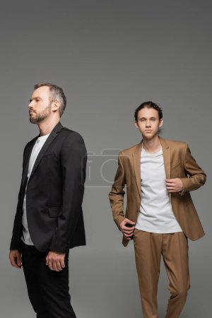 Photo for Well dressed father and confident teenage son in suits standing together isolated on grey - Royalty Free Image