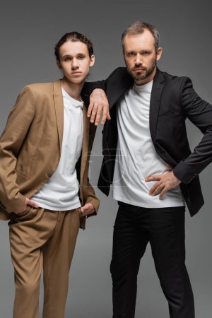confident father and teenage son in suits posing together isolated on grey 