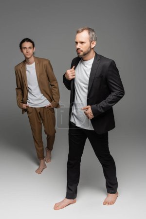 full length of well dressed man walking with bare feet next to stylish teenage boy on grey 