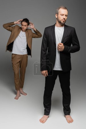full length of well dressed and barefoot man standing next to stylish teenage boy on grey 