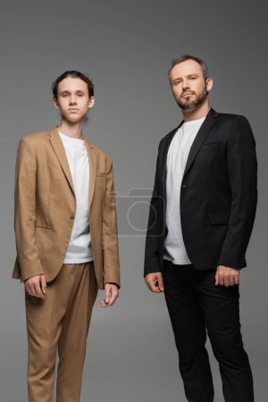 well dressed teenage boy and dad in suits looking at camera isolated on grey 