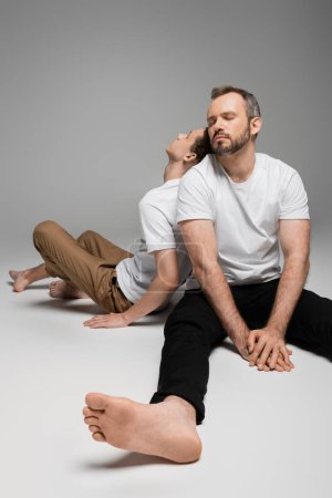 teenager boy leaning on back of bearded dad in white t-shirt while sitting on grey 