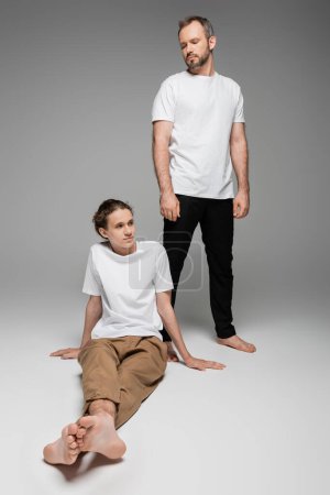 full length of father looking at his teenage son in white t-shirt on grey 