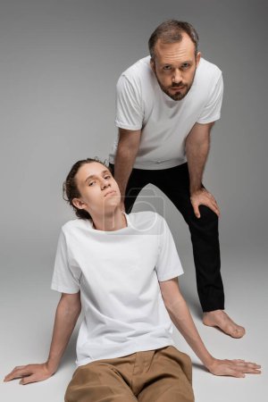 bearded father and teenage son in white t-shirts posing while looking at camera on grey 