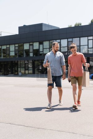 Bärtiger Vater und Teenager-Sohn laufen mit Einkaufstüten aus Einkaufszentrum 