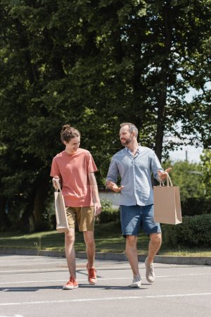 alegre papá y feliz hijo adolescente caminando con bolsas de compras al aire libre 