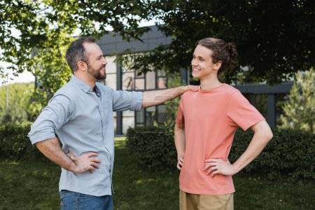 happy bearded father standing with hand on hip near teenage son in park 