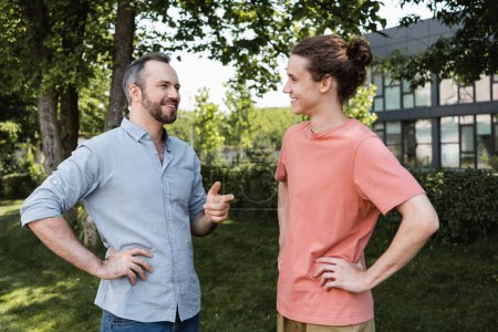 happy bearded father standing with hand on hip and pointing away near teenage son in park 