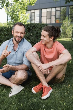 cheerful bearded father and teenage son sitting together on green lawn 