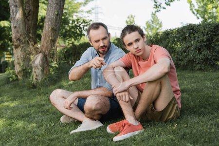 bearded man pointing away near teenager son while sitting together on green lawn 