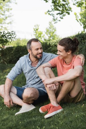 happy bearded father and cheerful teenage son sitting together on green lawn 