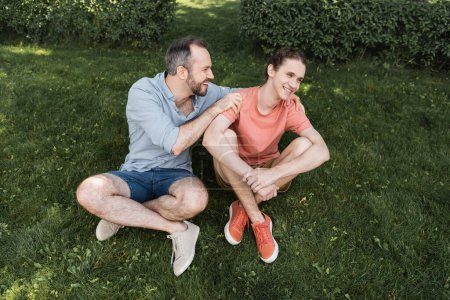 Blick von oben auf den glücklichen bärtigen Vater, der seinen Teenager-Sohn umarmt, während er zusammen auf dem grünen Rasen sitzt 