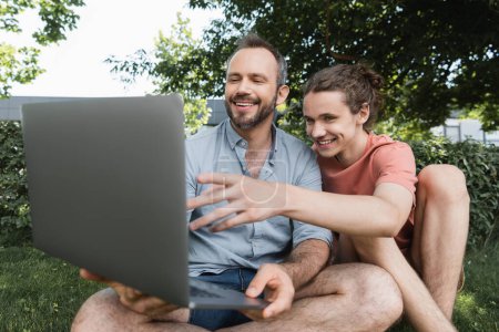 smiling father using laptop next to happy teenage boy while sitting together on green lawn 