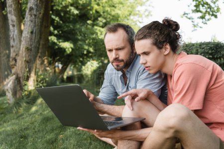 concentrated father and son looking at laptop while sitting together on green lawn 