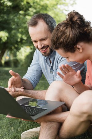 happy father pointing at laptop next to teenage boy while sitting together on green lawn 