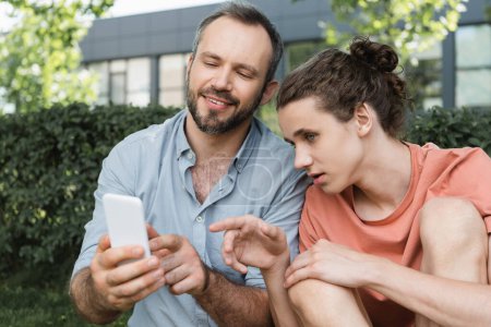 teenage boy pointing with finger at smartphone next to happy father in green park 