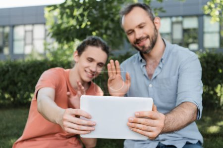 Téléchargez les photos : Joyeux papa et adolescent fils agitant les mains tout en ayant appel vidéo sur tablette numérique - en image libre de droit