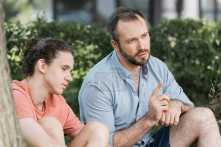 Foto de Padre barbudo apuntando mientras está sentado junto a su hijo adolescente y hablando en el parque verde - Imagen libre de derechos