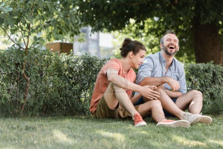glücklicher Vater lacht, während er neben pubertierendem Sohn auf grünem Rasen im Park sitzt 