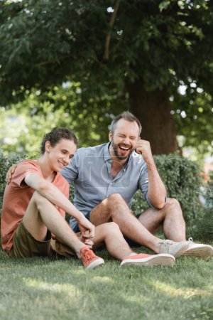 cheerful father and son sitting in summer clothes on green lawn in park 