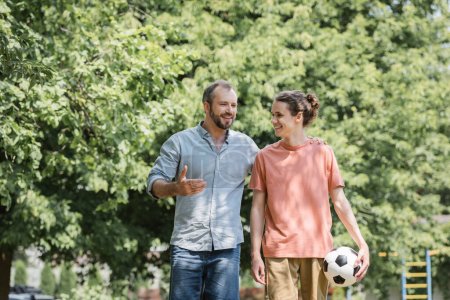 Téléchargez les photos : Heureux père étreignant adolescent fils tenant football tout en marchant dans vert parc - en image libre de droit