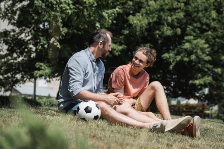 happy teenage boy sitting near soccer ball with father in green summer park 