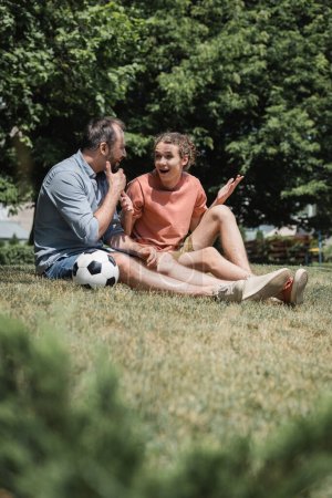 amazed teenage boy sitting near soccer ball with father in green summer park 