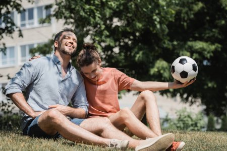 teenage boy sitting near joyful father and holding soccer ball in green summer park 