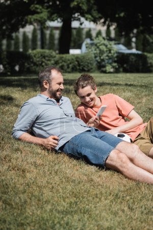 bearded man smiling while using smartphone and resting on lawn near teenage son after playing football 