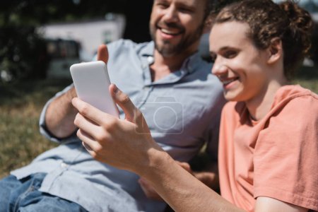 bearded father and happy teenage son smiling while looking at smartphone 