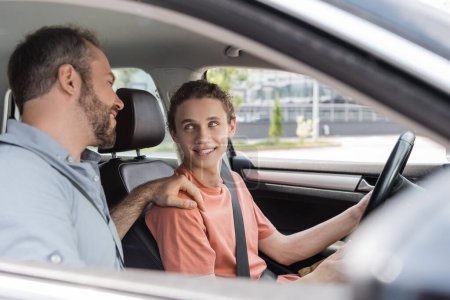 cheerful father putting hand on shoulder of teenage son while teaching him how to drive car 