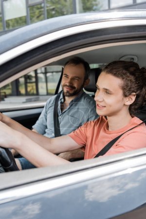happy teenage boy holding steering wheel while driving car next to dad 