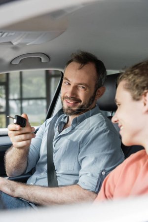 cheerful and bearded man holding car key next to son in modern auto 