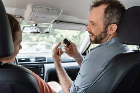 cheerful and bearded dad pointing at car key next to son in modern auto 